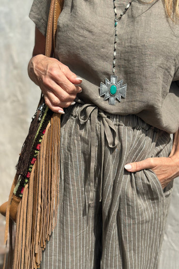 Person wearing a gray shirt and striped pants with a fringe bag and decorative necklace.