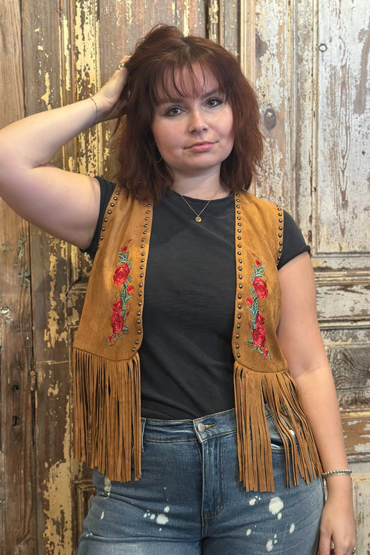 Person wearing a brown fringe vest with floral embroidery against a rustic wooden background