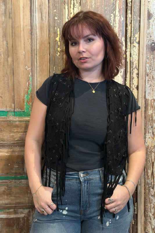 Woman wearing a black fringe vest over a dark top and jeans, standing against a wooden background.
