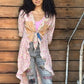 Woman standing against a wooden wall holding a drink, wearing a pink cardigan and ripped jeans.