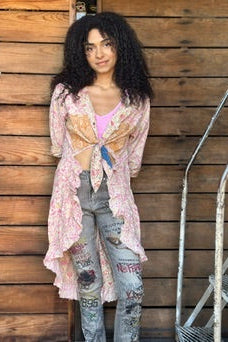 Woman standing against a wooden wall holding a drink, wearing a pink cardigan and ripped jeans.