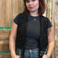Woman wearing a black fringe vest over a dark top and jeans, standing against a wooden background.