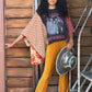 Woman wearing a colorful outfit with a poncho and yellow pants, standing against a wooden wall.