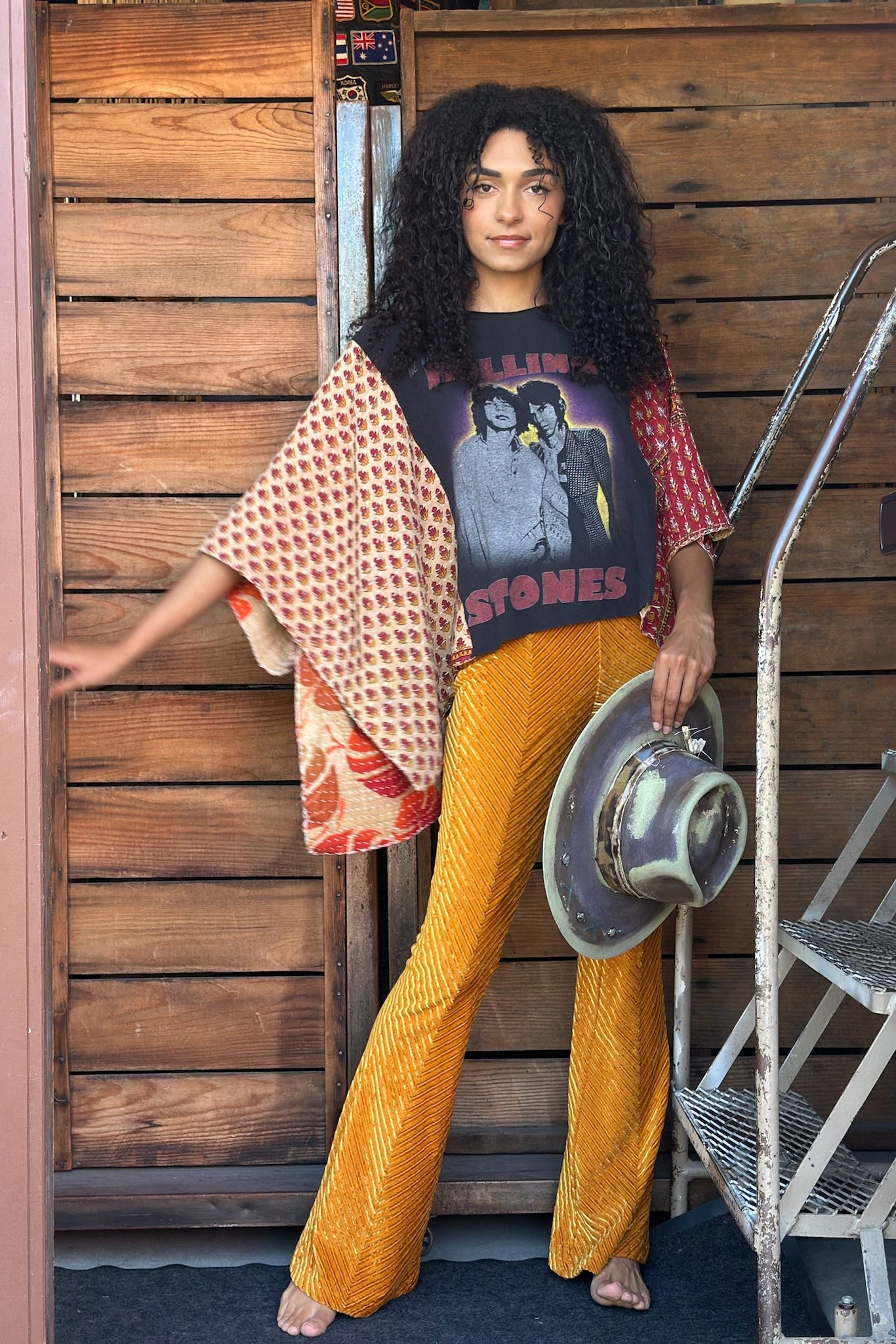 Woman wearing a colorful outfit with a poncho and yellow pants, standing against a wooden wall.