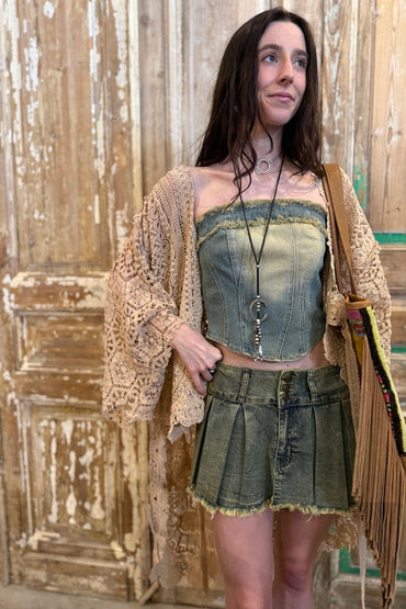 Woman wearing a denim corset top and shorts with a beige cardigan against a rustic wooden wall.