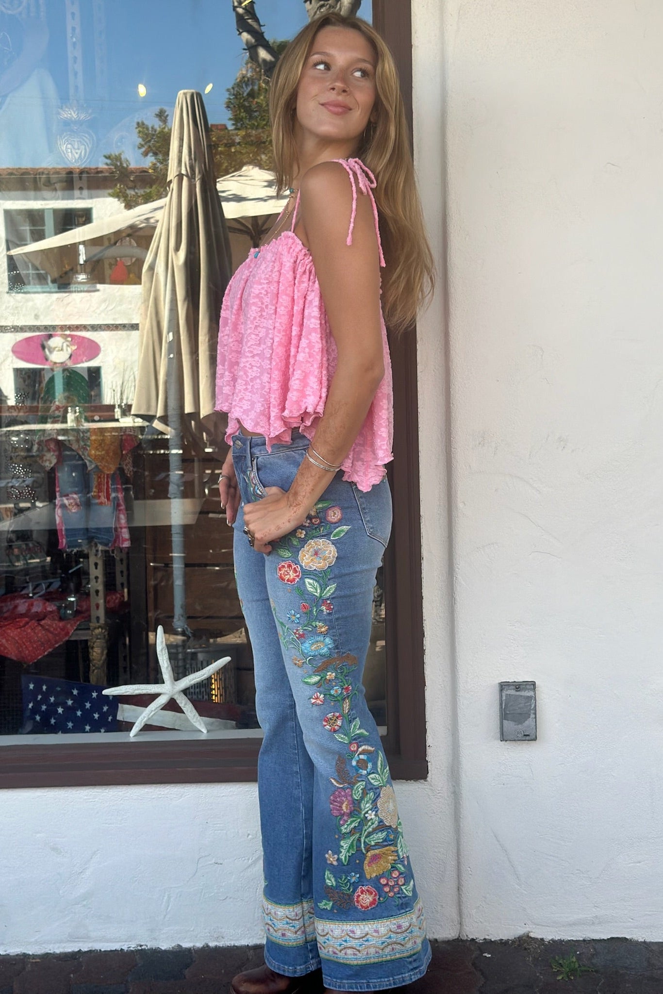 Woman wearing a pink top and floral jeans standing in front of a store window.