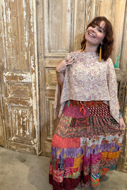 Woman wearing a floral top and colorful patchwork skirt standing in front of a wooden wall.