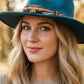 Woman wearing a teal hat with a brown band in a forest setting