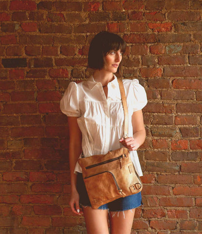 Model wearing a Trestles Clothing Company Aiken convertible brown leather bag with zip pockets against a brick wall