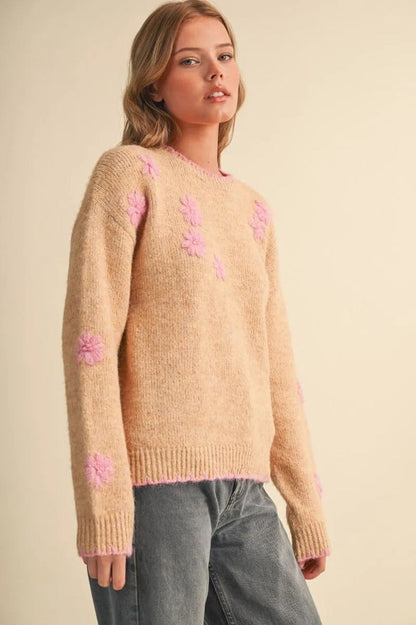 Model wearing a Trestles Clothing Company beige knit sweater with pink floral embroidery and scalloped contrast edges.