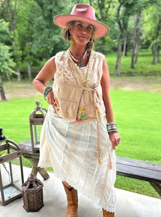 Trestles Clothing Company beige wrap vest with floral patches and lace, worn over a white dress with a pink hat.