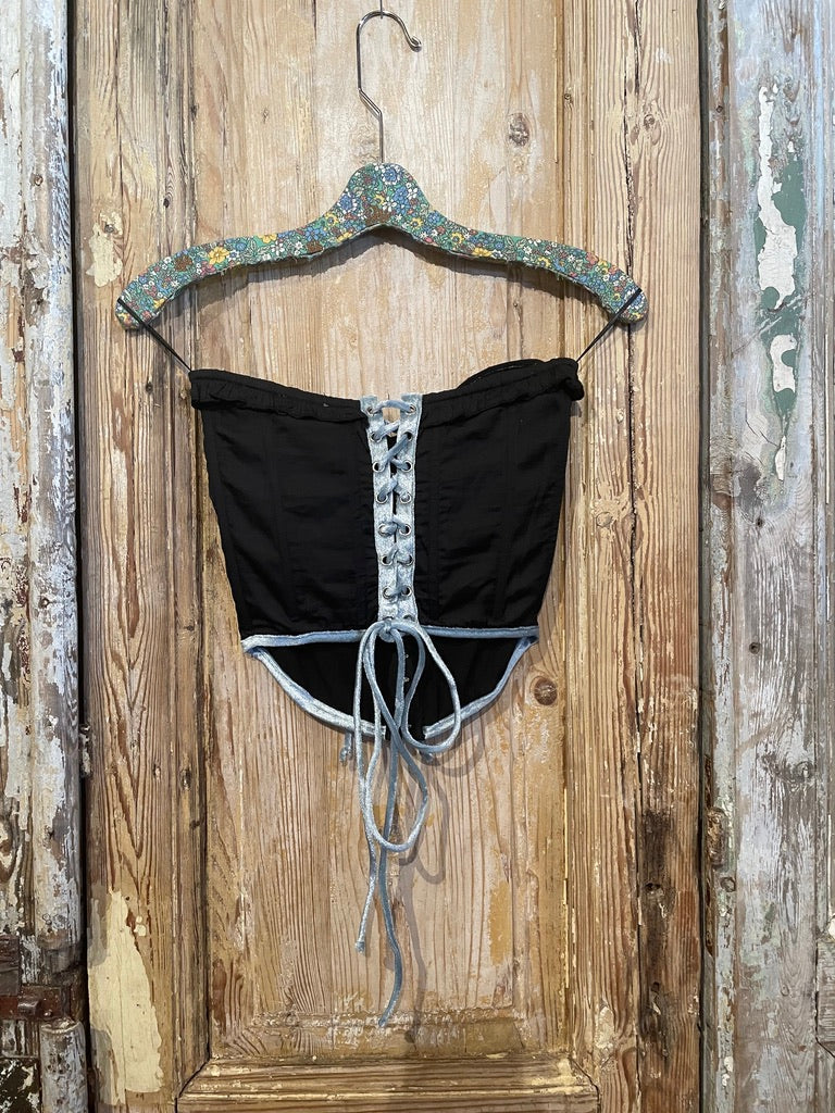 Trestles Clothing Company black corset top with blue velvet trim and front lace-up detail hanging on a hanger.