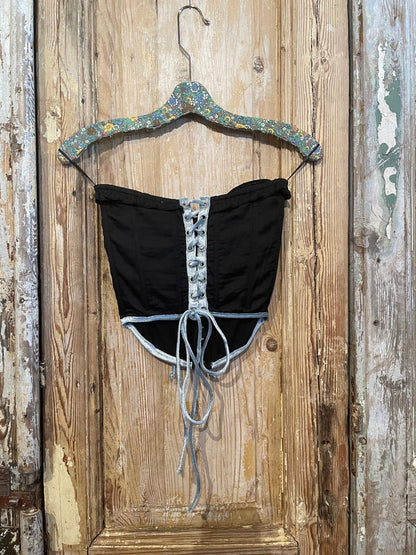 Trestles Clothing Company black corset top with blue velvet trim and front lace-up detail hanging on a hanger.