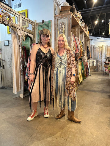 Two women wearing Trestles Clothing Company crochet fringe vests in a vintage-inspired boutique setting.