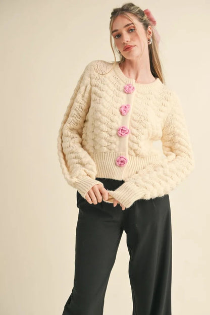 Trestles Clothing Company ivory knit cardigan with pink rosette buttons, long puff sleeves, and a cropped fit.