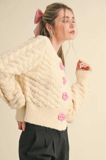 Trestles Clothing Company ivory knit cardigan with pink rosette buttons, puff sleeves, and a cropped fit.