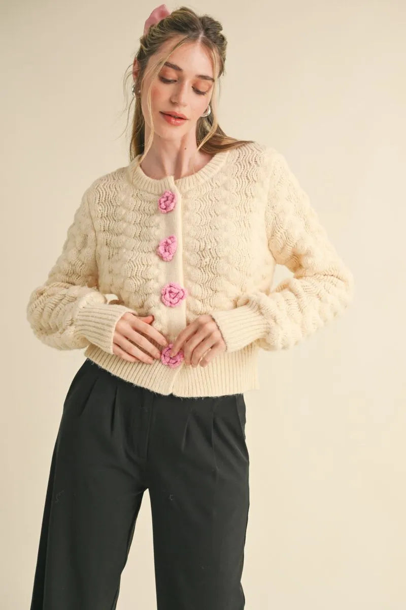Trestles Clothing Company ivory knit cardigan with pink rosette buttons, long sleeves, and a cropped fit.