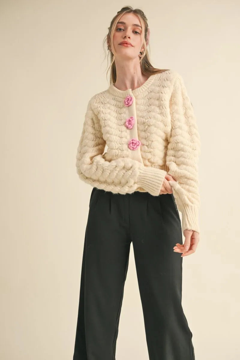 Trestles Clothing Company ivory knit cardigan with pink rosette buttons, long sleeves, and a cropped fit on a model.