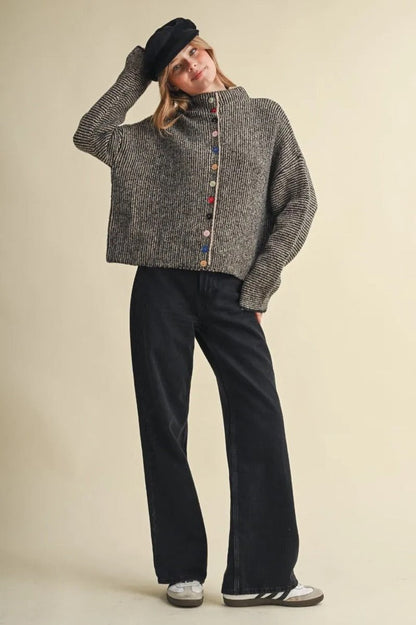 Model wearing a Trestles Clothing Company grey knit sweater with colorful mismatched buttons and a black beret