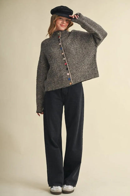 Model wearing a Trestles Clothing Company grey ribbed knit sweater with mismatched colorful buttons and black jeans.